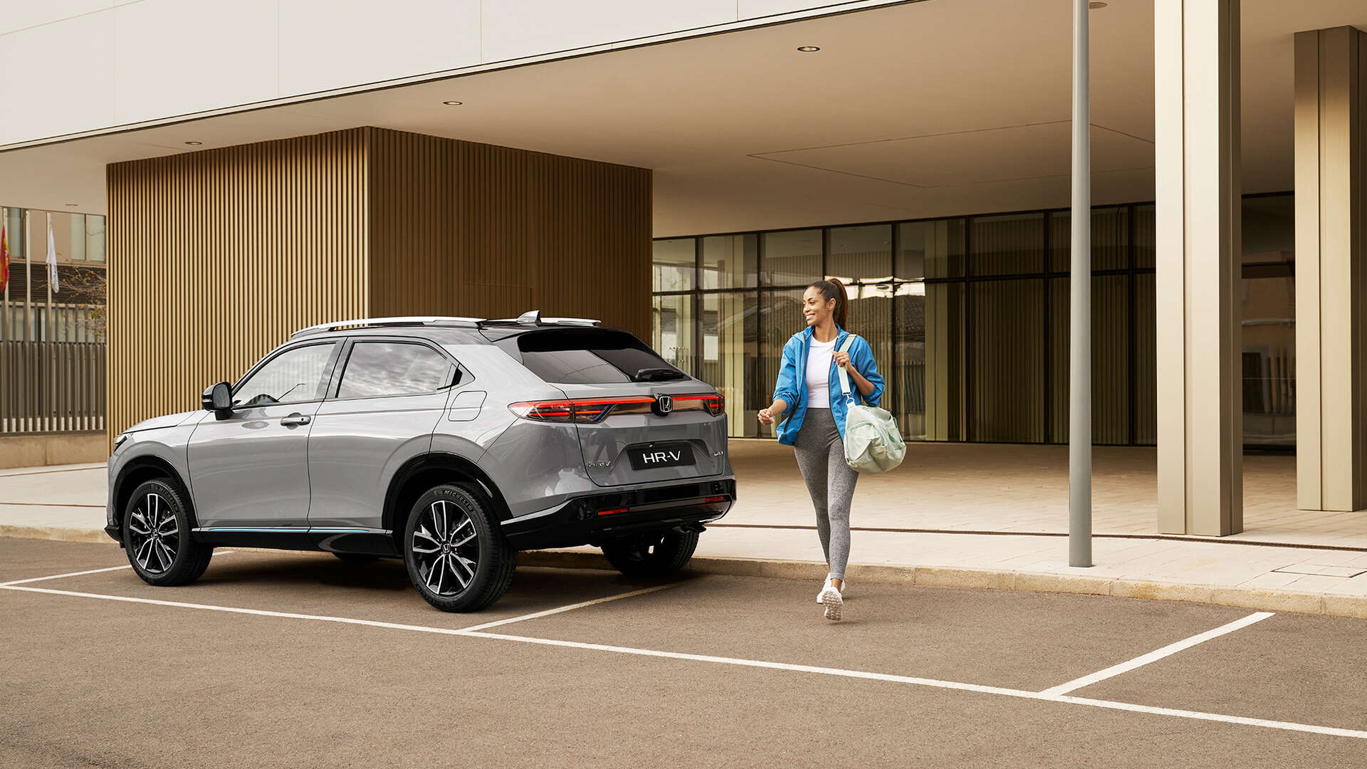 Strieborná Honda HR-V zaparkovaná pred modernou budovou, zatiaľ čo okolo nej prechádza žena s taškou, čo naznačuje každodenné mestské využitie a komfort.